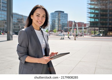 Young happy Asian business woman entrepreneur wearing suit holding digital tablet standing in big city on busy street, using smart business software tech for online work on pad computer outdoor. - Powered by Shutterstock