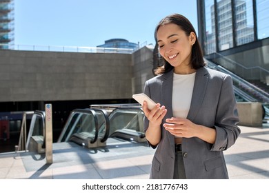 Young happy Asian business woman wearing suit holding mobile phone standing in city subway using smartphone for texting, checking apps for public transport, metro or travel guide. - Shutterstock ID 2181776743