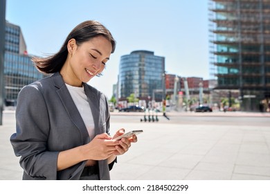 Young happy Asian business woman wearing suit holding mobile phone standing in big city on busy downtown street, smiling lady using smartphone for texting, using apps outside. - Shutterstock ID 2184502299