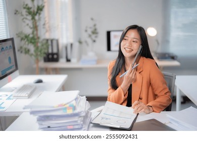 Young happy asian businesswoman holding a pen and talking on mobile phone while sitting at desk and analyzing financial charts in modern office - Powered by Shutterstock