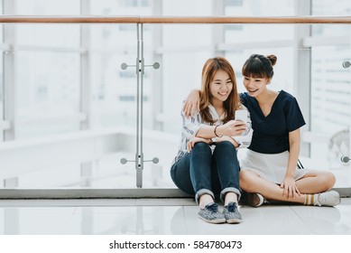 Young Happy Asian Girls Best Friends Laugh And Smile While Sitting And Having Fun With Smart Phone Mobile Indoor.