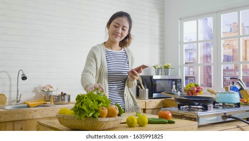 Young Happy Asian Japanese Smiling Woman Wear Sweater Looking Recipe In Mobile Phone While Standing In Kitchen. Vegetable Salad Dieting Healthy Lifestyle Concept. Girl Cooking At Home Prepare Food