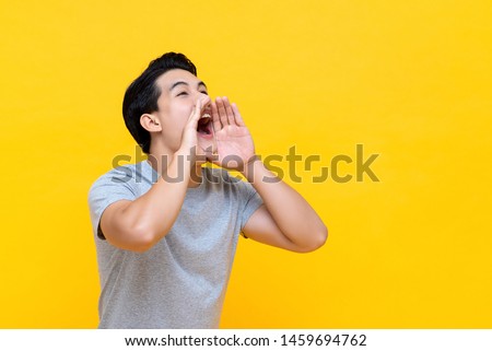 Young happy asian man announcing information with hands cup around mouth isolated on yellow studio background