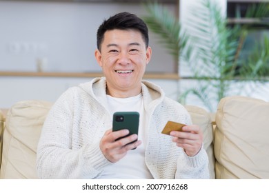 Young Happy Asian Man Shopping Online In Internet Store Using Mobile Phone And Credit Card. Male Enters Data Of Paying For Services Or Tickets In The Application Sitting On Couch At Home. Easy App