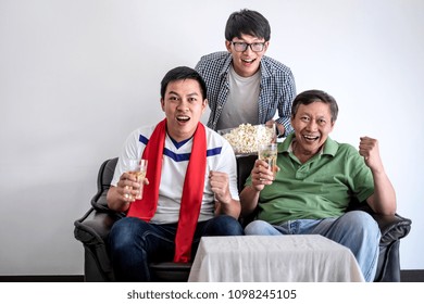 Young Happy Asian Men Family Or Football Fans Watching Soccer Match On Tv And Cheering Football Team, Celebrating With Drink Beer And Eat Popcorn At Home, Sports And Entertainment Concept.