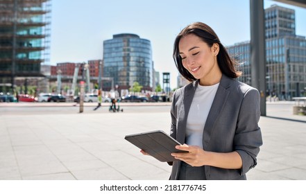 Young Happy Asian Professional Woman Wearing Suit Holding Digital Tablet Standing In Big City On Busy Street, Smiling Lady Using Smart Business Software For Online Work On Pad Computer Outside.