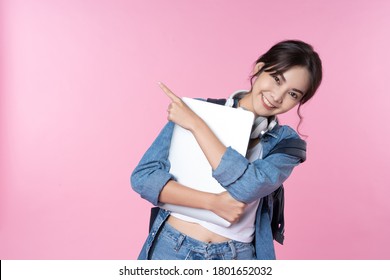 Young happy Asian woman holding laptop pointing finger hands up to copy space she wear jean jacket and carry backpack shirt shoot in isolated on pink background