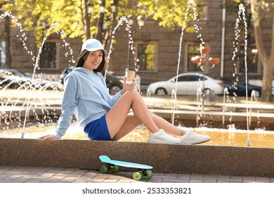 Young happy Asian woman in stylish white cap with skateboard and cup of coffee sitting near fountain on city street - Powered by Shutterstock