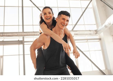 young happy athletic couple in sportswear runs and rejoices in bright gym, the guy carries the girl on his back and smiles, the concept of freedom - Powered by Shutterstock