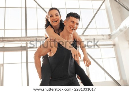young happy athletic couple in sportswear runs and rejoices in bright gym, the guy carries the girl on his back and smiles, the concept of freedom