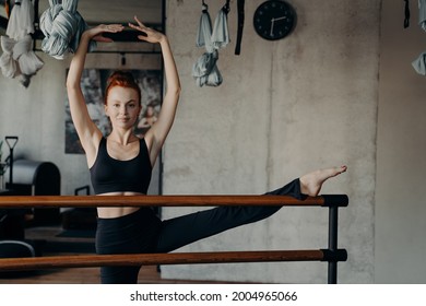 Young happy attractive redhaired woman in active wear standing in fitness studio with one leg on ballet barre, having hands above head in classical ballet arm position looking into camera
