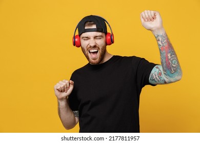 Young happy bearded tattooed man 20s he wears casual black t-shirt cap headphones listen to music dance have fun in free time isolated on plain yellow wall background studio. People lifestyle concept
