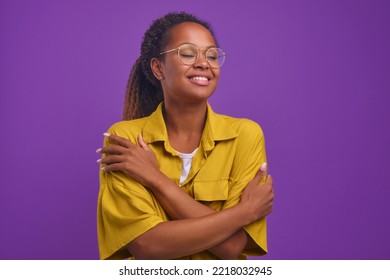 Young happy beautiful African American woman millennial with curly hair hugs herself to cheer up and get rid of loneliness dressed in casual yellow shirt, stands on isolated purple background.