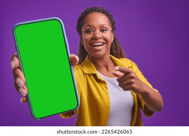 Young happy beautiful African American woman demonstrates phone with green display admiring new online shopping website or interesting news article stands in purple studio. Smartphone, applications - Powered by Shutterstock