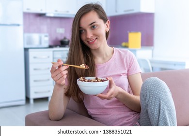 Young, Happy Beautiful Brunette Woman In Pajamas Eating Chocolate Balls For Cereals Breakfast While Watching Tv At Home 