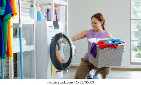 Young happy beautiful friendly asian female long brown ponytail hair housekeeper wear purple sleeveless shirt and brown pants put dirty clothes from basket into white washing machine in laundry room.