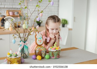 A young happy beautiful girl with 2 braids in peach colored dress is sitting in the kitchen with Easter cakes, colorful eggs and tree branches for Easter home decoration. Kitchen in the background