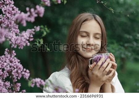 Young happy beautiful girl with long hair with smile and closed eyes holding transparent cup with pink flowers in her hands in garden or park, blooming branches, idea of relax and rest with nature