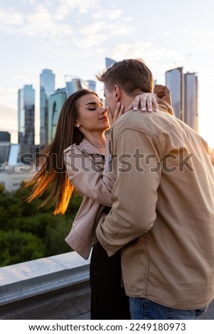 Young happy beautiful loving couple on a surprise romantic date on a rooftop on a Saint Valentine's Day. Attractive man and woman, male and female hugging, kissing. Sunset, skyscrapers urban view