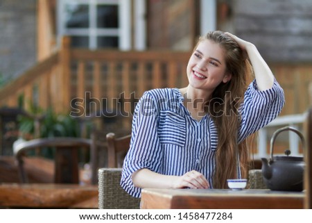 Young happy,  beautiful, smiling girl,  sitting at the table on the chair at the Asian style cafe,  drinking tea from authentic Chinese teapot against blurred background 