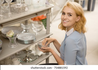 Young happy beautiful woman smiling to the camera over her shoulder while choosing dinnerware at the local houseware store copyspace dishes plates dining room decor purchasing consumerism