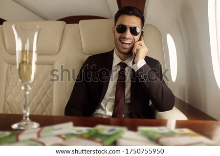 Young happy billionaire on board of his private jet sitting at table with champagne and euros in cash, talking on phone and laughing happily, enjoying financial success