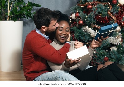 Young happy biracial couple sitting at home in front of the Christmas tree decorated with decorations exchanging gifts for the New Year looking forward to the festive mood - Powered by Shutterstock