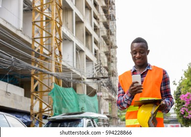 Young happy black African man construction worker smiling and using mobile phone while holding safety helmet and clipboard at building site