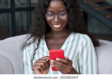 Young happy black African woman wearing glasses using mobile digital apps on cell phone tech device for online shopping or dating sitting on sofa at home, texting on smartphone, writing social post.
