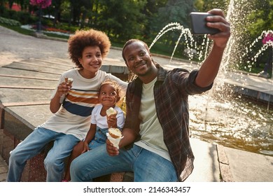 Young Happy Black Family Eating Ice Cream Cones And Taking Selfie On Mobile Phones Near Fountain In Park. Family Relationship And Enjoying Time Together. Fatherhood And Parenting. Sunny Summer Day