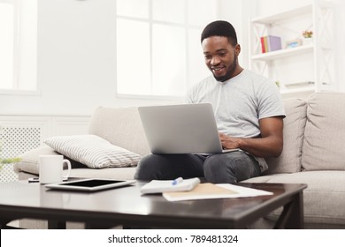 Young Happy Black Man Chatting Online On Laptop With Friend At Home. Casual Guy Sitting On Beige Couch In Light Livingroom, Copy Space