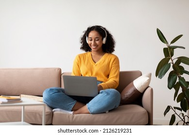 Young Happy Black Woman In Headphones Using Laptop Pc, Working Online, Sitting On Couch At Home, Copy Space. Millennial African American Female Having Remote Meeting, Speaking On Webcam