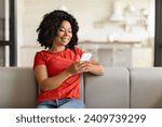 Young happy black woman texting on her phone at home, smiling african american lady in red t-shirt comfortably seated on couch, engrossed in messaging on smartphone in living room interior