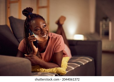 Young Happy Black Woman Using Smart Phone And Making A Phone Call While Relaxing On The Sofa At Night At Home. 