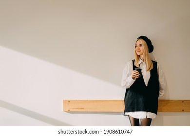 Young happy blonde woman standing at a white wall, holding a cup of coffee, smiling.