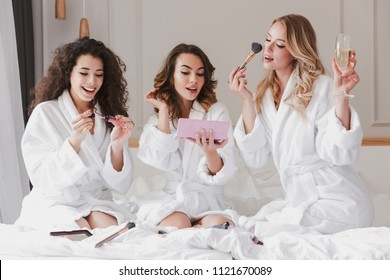 Young happy bride and bridesmaids 20s wearing housecoat drinking champagne and applying makeup during girls party while resting in luxury bedroom at hotel room