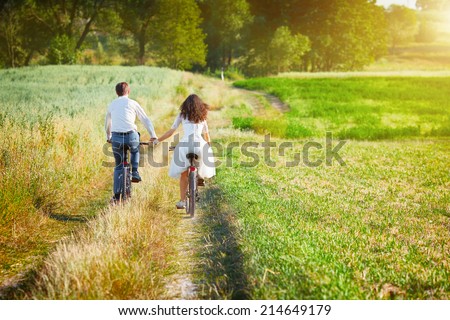 Young happy bride and groom ride bicycles in the meadow back to camera and holding hands.