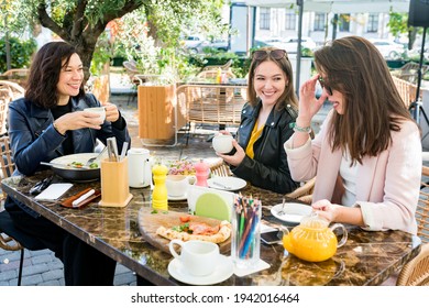 Young Happy Brunette Women Friends In Casual Clothes Having Fun And Laughing During Brunch In Cafe Outside On Sunny Day. Selective Focus