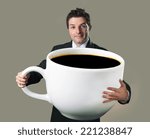 young happy business man holding a funny huge and oversized cup of black coffee in caffeine addiction concept isolated on even background