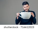 young happy business man holding a funny huge and oversized cup of black coffee in caffeine addiction concept isolated on even background