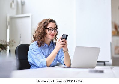 Young happy business woman, smiling beautiful professional lady worker looking at smartphone using cellphone mobile cellular tech working at home or in office checking cell phone sitting at desk. - Powered by Shutterstock