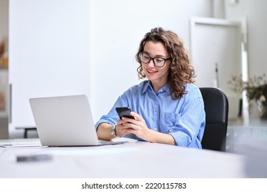Young happy business woman, smiling pretty professional businesswoman worker looking at smartphone using cellphone mobile technology working at home or in office checking cell phone sitting at desk. - Shutterstock ID 2220115783