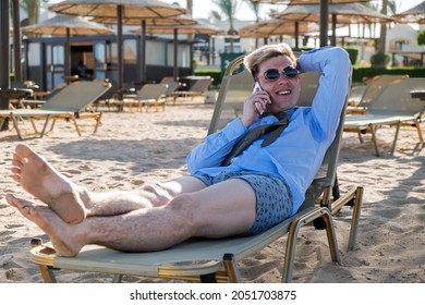 Young Happy Businessman Downshifter Talks On The Phone Lying On A Sun Lounger In The Middle Of The Beach In A Shirt, Tie And Boxer Shorts