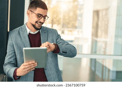 Young happy businessman with reading glasses checking time on his watch while holding digital tablet outside modern work office. - Powered by Shutterstock