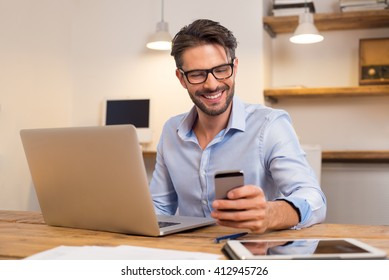 Young happy businessman smiling while reading his smartphone. Portrait of smiling business man reading message with smartphone in office. Man working at his desk at office.