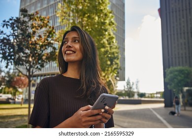 Young happy businesswoman standing in sun smiling and holding mobile phone - Shutterstock ID 2165595329