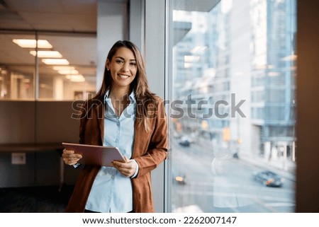 Photo of Young happy businesswoman using digital tablet while standing by the window in the office and looking at camera. Copy space. 