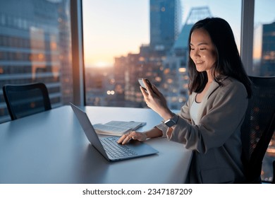 Young happy busy Asian business woman executive using laptop mobile cell phone tech at night in dark office. Professional businesswoman holding smartphone, working on cellphone with evening city view. - Powered by Shutterstock