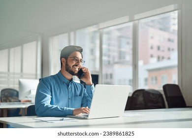 Young happy busy latin businessman talking on phone using laptop computer in office. Smiling hispanic business man making call on mobile cellphone, consulting client or having work cell conversation. - Powered by Shutterstock