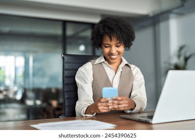Young happy busy professional young African American business woman entrepreneur holding cellphone using mobile looking at cell phone tech sitting at work desk in office working on laptop.